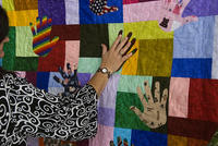 A quilt representing diversity. A female hand touching the quilt. The quilt consists of multiple colourful patches, some of the palm-shaped. The woman's palm is open against one of the palms on the quilt. 