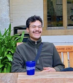Photo of student Batelaan sitting relaxed and smiling, in a garden pub, blue cup on the table in front of him.